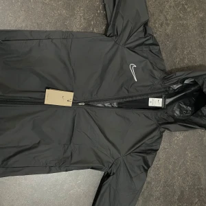 Svart regnjacka från Nike med huva - Svart Nike regnjacka med huva och dragkedja framtill. Jackan har en clean look med diskret Nike-logga på bröstet och är tillverkad i lätt syntetmaterial. Perfekt för blåsiga dagar och regniga dagar även enkel att slänga på sig när vädret svänger.