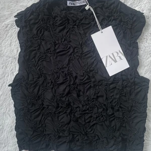 Svart ruched topp från Zara - Cool svart topp från Zara med ruched effekt över hela tyget som ger en unik struktur. Ärmlös modell med hög hals och croppad passform. Perfekt att styla med jeans eller kjol för en trendig look. Pris kan diskuteras 