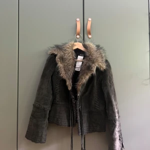 Grå läder och pälsjacka från Morgan med snörning - Säljer en grå läder pälsjacka från Morgan med riktigt päls runt kragen och coola snörningsdetaljer längs sidorna. Jackan är gjord i läder, har en cropat fit med långa ärmar. 