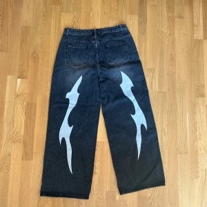 Wide jeans med tryck i svart denim - Säljer ett par svarta wide jeans med coolt vitt tryck på baksidan av benen. Materialet är robust denim som ger en snygg och avslappnad look.