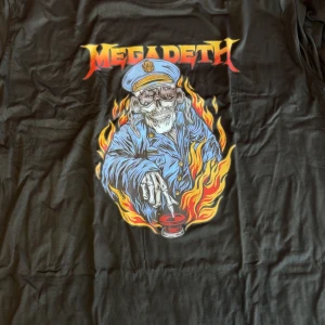 Megadeth svart t-shirt med tryck - Svart t-shirt med stort Megadeth-tryck på bröstet. Motivet visar en skelettfigur i blå uniform omgiven av eldflammor och med ett rött knapptryck. Perfekt för dig som gillar metal och vill sticka ut med en cool bandtröja.