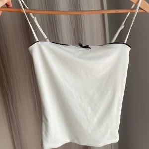 Vit linnetopp med svart kant från BikBok - Supersöt vit linnetopp från BikBok i storlek XS. Toppen har tunna axelband, svart spetskant upptill och en liten svart rosett framtill. Perfekt för sommardagar eller att styla lager på lager. Materialet känns mjukt och lätt mot huden.