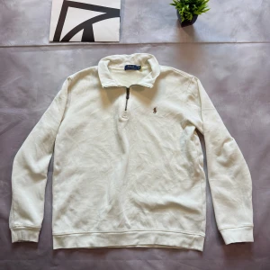 Vit half zip tröja från Polo Ralph Lauren - Vit långärmad tröja från Polo Ralph Lauren med klassisk krage och half zip-dragkedja. Tröjan har broderad logga på bröstet och är tillverkad i mjuk bomull. Perfekt för en clean och stilren look.