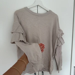 Beige volangtröja från Gina Tricot - Supersöt beige tröja från Gina Tricot med långa ärmar och volangdetaljer på ärmarna. Tröjan är i mjuk bomullsmix och har en relaxed fit. Perfekt för dig som gillar en chill men ändå trendig stil.