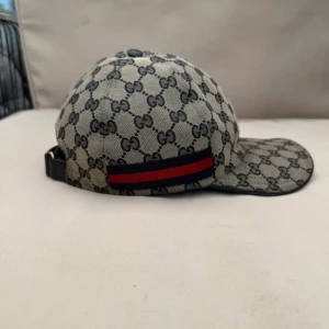 Gucci keps med GG-mönster och röd-blå rand - Snygg keps från Gucci med klassiskt GG-monogram i svart på beige botten. Kepsen har justerbar rem bak i svart skinn och en ikonisk röd-blå rand på sidan. Tillverkad i bomullsmix med detaljer i skinn. Perfekt för dig som vill sticka ut med lyxig streetstyle.