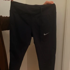Nike- Svarta  Dri-Fit träningsbyxor - Svarta träningsbyxor från Nike med Dri-Fit-teknologi. Byxorna har en tight passform, snörning i midjan och en diskret Nike-logga på benet. Perfekta för dig som vill ha bekväma och funktionella byxor till träningen. Petite M