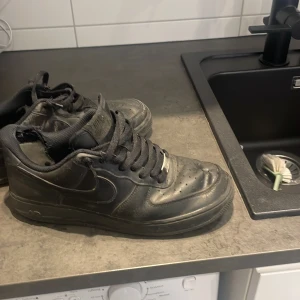 Svarta Nike Air Force 1 sneakers - Klassiska svarta Nike Air Force 1 sneakers i skinn med rund tå och snörning. Snygg streetstil med perforerade detaljer på tån och ikonisk Nike-logga på sidan. Perfekta för dig som gillar en tidlös och urban look.