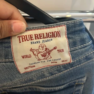 True Religion bootcut jeans blå - Säljer ett par snygga blå jeans från True Religion med klassiska kontrastsömmar och ikonisk logga på bakfickan. Modellen har bootcut-ben och låg midja, perfekt för dig som gillar en avslappnad men ändå trendig look. Jeansen är i klassiskt denim med coola detaljer.