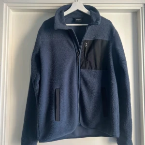 Blå fleecejacka från Everest M - Mysig blå fleecejacka från Everest med svarta detaljer och dragkedja framtill. Jackan har en hög krage, två sidofickor och en bröstficka med dragkedja. Perfekt för kyliga dagar och outdoor vibes.