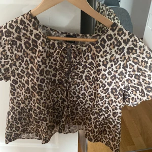 Leopardmönstrad blus från Gina Tricot - Säljer en kortärmad blus från Gina Tricot med leopardmönster i brunt, beige och svart. Toppen har snörning framtill och en luftig, lätt passform. Perfekt för dig som vill sticka ut med ett djurmönstrat plagg. Helt nytt