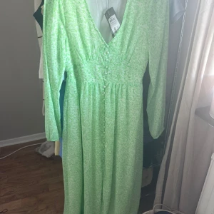 Grön långklänning från ONLY - Ljusgrön långklänning från ONLY med v-ringning, långa ärmar och knappar framtill. Klänningen har ett diskret mönster i vitt och grönt och är tillverkad i ett lätt och flowy material. Perfekt för dig som gillar en feminin och trendig stil.