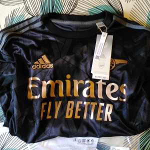 Arsenal Saka #7 svart bortatröja XS - Svart Arsenal bortatröja från Adidas med Saka nummer 7 på ryggen. Tröjan har guldiga detaljer, Adidas-logga och klubbmärke broderat, samt Emirates-tryck på bröstet. Tillverkad i lätt och ventilerande polyester med geometriskt mönster. Perfekt för dig som älskar fotboll och Arsenal. #arsenal #fotboll