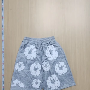 Denim tears  - Snygga grå shorts med elastisk midja och snörning. De har ett unikt vitt blommigt mönster över hela plagget och är tillverkade i mjuk bomull. Perfekta för en chill sommardag eller när du vill ha en avslappnad look. Kommer med t-shirt också.