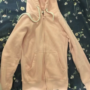 Ljusrosa hoodie från Russedress XS - Säljer en ljusrosa hoodie från Russedress i storlek XS. Tröjan har dragkedja, huva med snörning och två stora fickor framtill. Mjuk insida och klassisk passform, perfekt för chill dagar. Diskret Russedress-logga på ärmen.