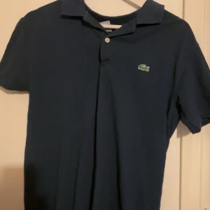 2-pack Lacoste pikétröjor marin & beige - Två klassiska Lacoste pikétröjor i marinblått och beige. Båda har den ikoniska gröna krokodilloggan på bröstet, krage och knappslå. Perfekt för dig som gillar stilren och tidlös design. Tillverkade i mjuk bomull och med kort ärm för en chill vibe.