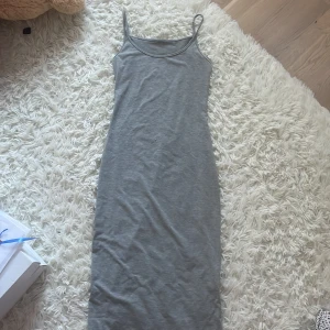 Grå långklänning med smala axelband - En enkel och stilren grå långklänning med smala axelband och rak passform. Klänningen är ärmlös och gjord i mjukt bomullstyg, perfekt för en chill och avslappnad look. Passar lika bra med sneakers som med sandaler.