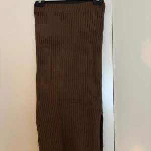 Brun ribbad långkjol från NA-KD - Säljer en brun ribbstickad långkjol från NA-KD i storlek XS. Kjolen har en snygg slits på sidan och är i mjukt stickat material som sitter skönt och följsamt. Perfekt för dig som gillar en clean och chill stil.
