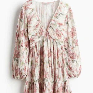 Blommig plisserad klänning med v-ringning - Superfin blommig klänning med plisserat tyg och v-ringad hals. Långa puffärmar och flowig passform ger en romantisk vibe. Basen är vit med rosa och gröna blommor, perfekt för vår och sommar.