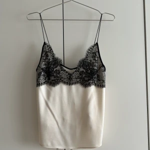 Vit sidenliknande topp med spets från Zara - Supersnygg vit sidenliknande topp från Zara i satin med tunna axelband och svart spetsdetalj upptill. Toppen har en lätt glansig finish och spetsen har blommigt mönster som ger en lyxig känsla. Perfekt att styla med jeans eller kjol för en trendig look.