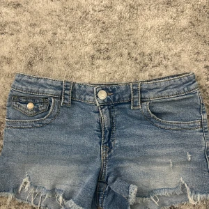Blåa jeansshorts från Gina Tricot - Snygga blå jeansshorts från Gina Tricot med fransig kant och klassiska fickor både fram och bak. Shortsen har bälteshällor, knapp och dragkedja framtill samt coola detaljer på bakfickorna. Perfekta för varma dagar och en avslappnad stil. Säljs pågrund av ingen användning 