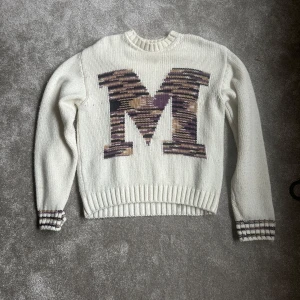 Vit stickad tröja med M från Missoni - Vit stickad tröja från Missoni med stor M i camo-liknande mönster på bröstet. Tröjan har ribbade muddar och nederkant, samt detaljer i brunt och lila på ärmsluten. Perfekt för dig som gillar statement-plagg och snygga detaljer.