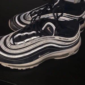 Nike Air Max 97 svart/vit sneakers - Nike Air Max 97 sneakers i svart och vitt med ikoniska vågformade linjer och reflekterande detaljer. Skorna har snörning, platt sula och synlig Air Max-dämpning i sulan. Perfekta för dig som gillar streetstyle och vill sticka ut.