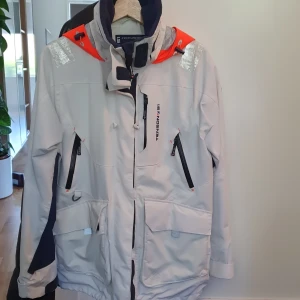 Tenson vindjacka med orange detaljer - Säljer en snygg vindjacka från Tenson i ljusgrått med mörkblå och orange detaljer. Jackan har flera dragkedjeförsedda fickor, hög krage och justerbar huva. Perfekt för blåsiga dagar och outdoor-äventyr. Stilsäker och funktionell design.