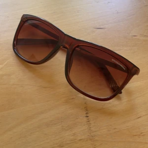 Bruna solglasögon från Dolce & Gabbana - Snygga solglasögon från Dolce & Gabbana med bruna, fyrkantiga bågar och matchande bruna glas. Klassisk och stilren design med diskret logga på sidan. Perfekta för soliga dagar och ger en cool vibe till din outfit.