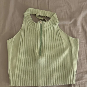 Ljusgrön ribbad halterneck topp - Superfin ljusgrön ribbad halterneck topp med dragkedja framtill. Toppen är ärmlös och har en tight passform med hög hals och öppen rygg. Perfekt för dig som gillar trendiga och stilrena plagg med snygga detaljer.