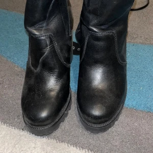 Svarta boots med klack och platåsula - Säljer ett par svarta boots i skinn med chunky klack och grov platåsula. Skorna har rund tå och markerade sömmar för en cool look. Perfekta för dig som vill ha lite extra höjd och attityd i outfiten.