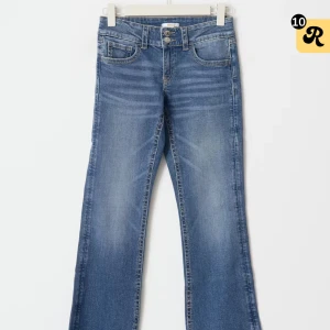Blå bootcut jeans med dubbla knappar - Snygga blå jeans med bootcut-modell och markerade vita sömmar. Byxorna har dubbla knappar i midjan, klassiska fem fickor och en cool tvättad look. Perfekta för dig som gillar retrovibbar och vill ha ett par jeans med lite extra attityd.