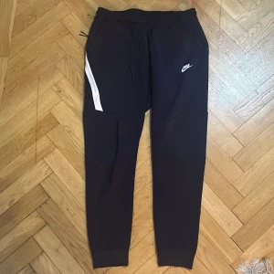 Svarta mjukisbyxor från Nike L - Snygga svarta mjukisbyxor från Nike i storlek L. Byxorna har en vit detalj på sidan, dragsko i midjan och klassisk Nike-logga på låret. Tillverkade i mjukt material med ribbade muddar vid bensluten för en skön passform.