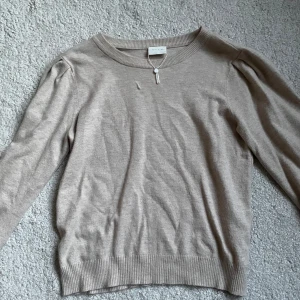 Beige stickad tröja från Vila - Supermjuk och skön beige stickad tröja från Vila med rund halsringning och diskreta puffärmar. Tröjan har ribbade muddar vid ärmslut och nederkant, vilket ger en snygg passform. Perfekt för alla tillfällen.