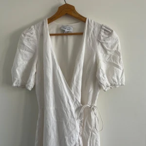 Vit omlottklänning från & Other Stories - Supersöt vit omlottklänning från & Other Stories med puffiga korta ärmar och knyt i midjan. Klänningen har v-ringad hals och volangkant nedtill, vilket ger en chill och feminin vibe. Tillverkad i linne, perfekt för varma dagar.