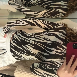 Zebramönstrad polotopp från Lindex - Säljer en långärmad polotopp från Lindex med svart och beige zebramönster. Toppens material är tunn och stretchig polyester, lite genomskinlig