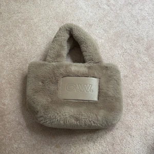 Beige fluffig handväska från C.W. - Supermjuk och fluffig beige handväska från C.W. med korta handtag och ett stort C.W.-märke framtill. Väskan är i fuskpäls och har ett rymligt innerfack med dragkedja. Perfekt för dig som vill ha en mysig och trendig accessoar till din outfit. Man kan ha ett långt band och även ta av det. Skriv om ni har några frågor 😊
