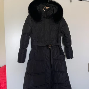 Svart lång dunjacka med fuskpäls - Lång svart dunjacka med fluffig fuskpäls runt huvan och bälte i midjan. Jackan är quiltad och har dragkedja framtill samt fickor med lock. Perfekt för kalla vinterdagar när du vill hålla stilen och värmen.helt ny.Eu storlek 40 märkt som XL