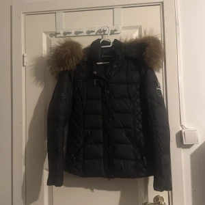 Svart pufferjacka Rockandblue med äkta  päls - Snygg svart pufferjacka från Rockandblue med stor fluffig päls på huvan. Jackan har quiltad design, dragkedjor på fickor och ärmar samt coolt Rockandblue-märke på ena armen. Perfekt för dig som vill ha en varm och trendig vinterjacka med edge.