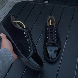 Svarta sneakers Lanvin - Stilrena svarta sneakers med ovandel i mocka och snygg lackad tå. Skorna har snörning och en platt sula, vilket ger en clean och modern look. Perfekta för dig som gillar en klassisk men ändå edgy stil.