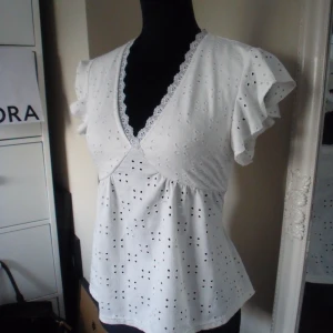 Vit blus med spets och volangärm - Supersöt vit blus med broderade hålmönster, v-ringning med spetskant och volangärmar. Luftig och feminin modell i bomull, perfekt för en fräsch och somrig look. Snyggt omlottliknande snitt över bysten och lätt utställd nertill.