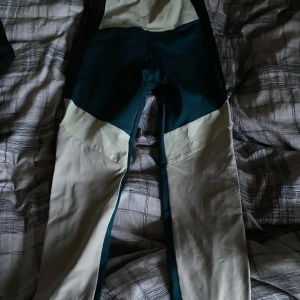 Röhnisch leggings XS i grönt och beige - Snygga leggings från Röhnisch i storlek XS. De har en cool färgblockad design i mörkgrönt och beige med logotyp längs benen. Materialet är stretchigt och passar perfekt för träning eller chill. Hög midja och tight passform för en sportig look.