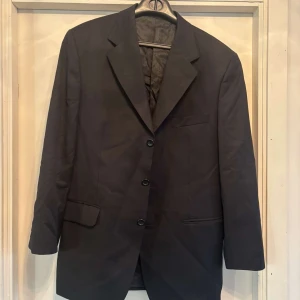 Svart kavaj från Oscar Jacobson - Klassisk svart kavaj från Oscar Jacobson med tre knappar framtill och snygga fickdetaljer. Rak passform och stilren design, perfekt för dig som gillar en tidlös look. Materialet är mjukt och känns lyxigt, med diskret mönstrad insida.