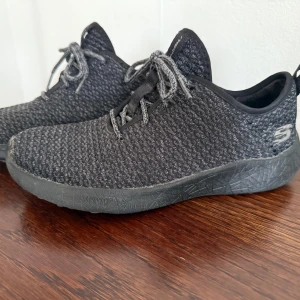  Skechers - Svarta sneakers från Skechers med stickad ovandel och matchande svarta snören. Skorna har en rund tå och platt sula med diskret mönster. Perfekta för dig som gillar en clean och sportig look. Snyggt S-logga på hälen.