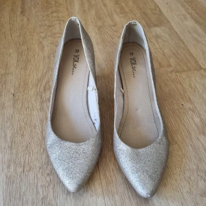 Glittriga guldiga pumps från VOX Shoes - Snygga pumps i glittrigt guld från VOX Shoes. Skorna har spetsig tå och låg klack, perfekta för att lyfta din outfit. De är täckta i glittrigt material som verkligen fångar ljuset och ger en festlig känsla.