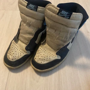 Nike Air Jordan 1 High blå/vit - Säljer ett par klassiska Nike Air Jordan 1 High i blått, vitt och svart skinn. Skorna har perforerad tå, svart swoosh och högt skaft. Perfekta för dig som gillar retrostil och streetwear. Ikonisk siluett och cool färgkombo.
