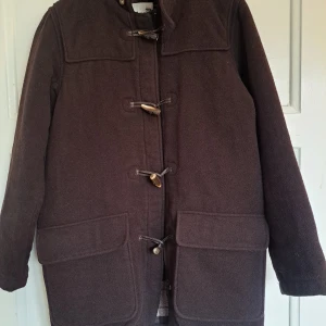 Brun duffeljacka från H&M, strl 158 - Mörkbrun duffeljacka från H&M med klassiska träknappar och stora fickor framtill. Jackan har rutigt innerfoder och är tillverkad i ullblandning. Perfekt för kyliga dagar och ger en tidlös look.