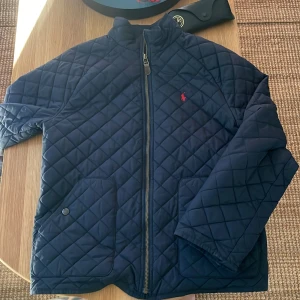 Mörkblå quiltad jacka Polo Ralph Lauren - Snygg mörkblå quiltad jacka från Polo Ralph Lauren med klassisk röd logga på bröstet. Jackan har dragkedja framtill, två stora fickor och hög krage. Perfekt för höst och vår, med stilren look och bekväm passform.