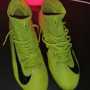Elite Nike Mercurial gula fotbollsskor - Snygga Nike Mercurial fotbollsskor i neon-gult med svarta detaljer och högt skaft. Skorna har snörning, svart swoosh på sidan och texten 'Mercurial' bak. Ovandelen är i syntetmaterial och designen är riktigt sportig och modern.