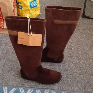 Bruna mockastövlar från Timberland - Helt nya. Snygga höga stövlar från Timberland i mörkbrun mocka med diskreta detaljer i ljusare brunt. Grov gummisula med Timberland-logga och präglad logga på skaftet. Stövlarna är vattentäta och har en klassisk, stilren look som passar till jeans eller kjol.