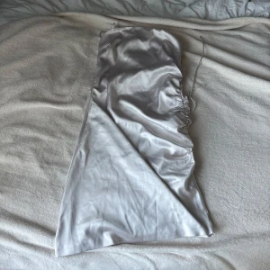 Ljus axelbandslös klänning NA-KD - Säljer en  axelbandslös kortklänning från NA-KD i glansigt satinliknande material. Klänningen har snörning upptill och en lätt rynkad detalj på sidan. Lätt snett skuren nertill. Dragkedja på sidan